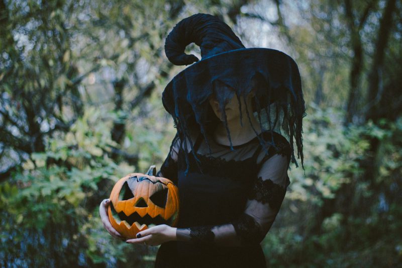 A witch in the forest holds a carved pumpkin