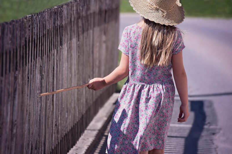 A little girl walks with a cane leaning against the fence